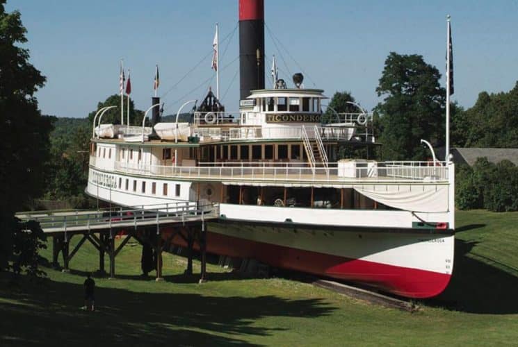 Antique steamboat named Ticonderoga beached on a wide grass lawn, accessible by a bridge.
