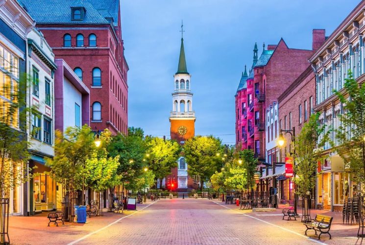 Looking down a brick street at dusk to a clock tower with a high spire, surrounded by a pretty downtown area.