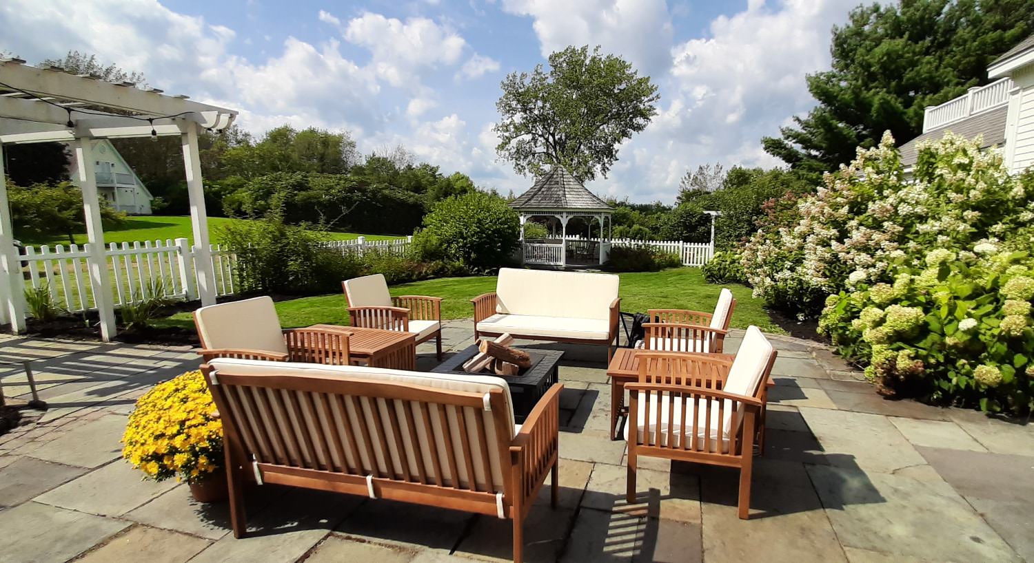 Light tan cushioned patio furniture on stone patio with green grass, flowering bushes, and vegetation in the background