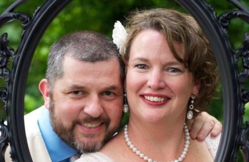 Bride wearing pearls and Groom in a blur shirt look through an ornate black picture frame.