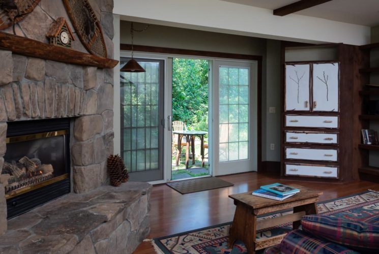 Large bedroom with a beautiful stone fireplace, plaid sofa, table and wooden floor looking out to patio through sliding door.