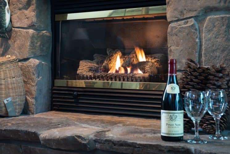 Bottle of red wine with two glasses and fishing decor on stone hearth in front of a fireplace burning merrily.