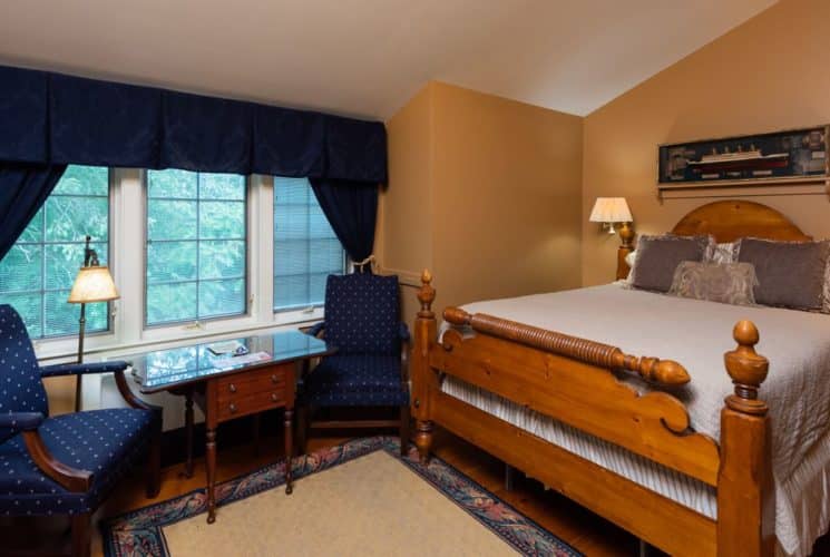 Bedroom decorated in tan and blue with a large wooden bed, blue chairs, side table and a large window.