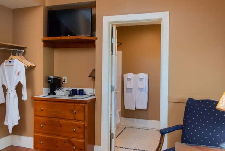 Clean and bright bedroom with tan walls, dresser and TV, white robe on hanger and blue chair next to a lamp.