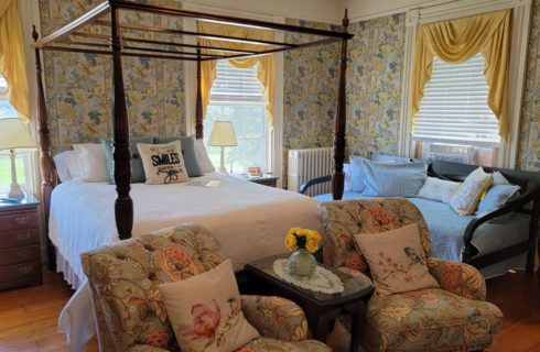 Empire-style 4-post bed made up in white in elegantly papered bedroom with armchair seating, a daybed and two bright windows.