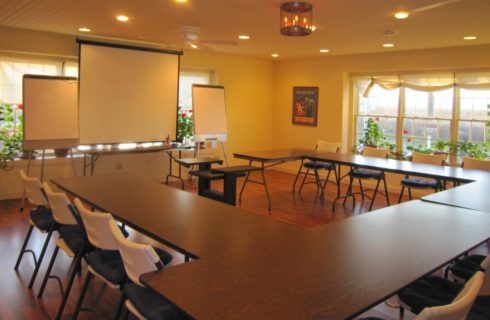 Meeting room set up with long tables and chairs with a screen and notepads at front.