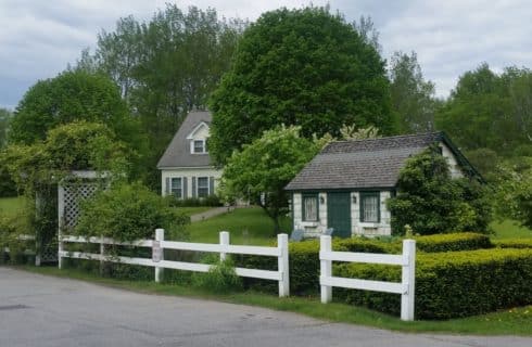 Pretty little white cottage with green door and shutters surrounded by a white wooden fence, backed by a house with a sloped roof.