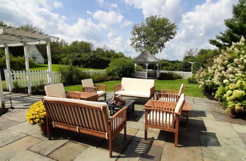 Stone patio on a cloudy/sunny day with wooden patio furniture, clouds of white flowering bushes, and a white gazebo.