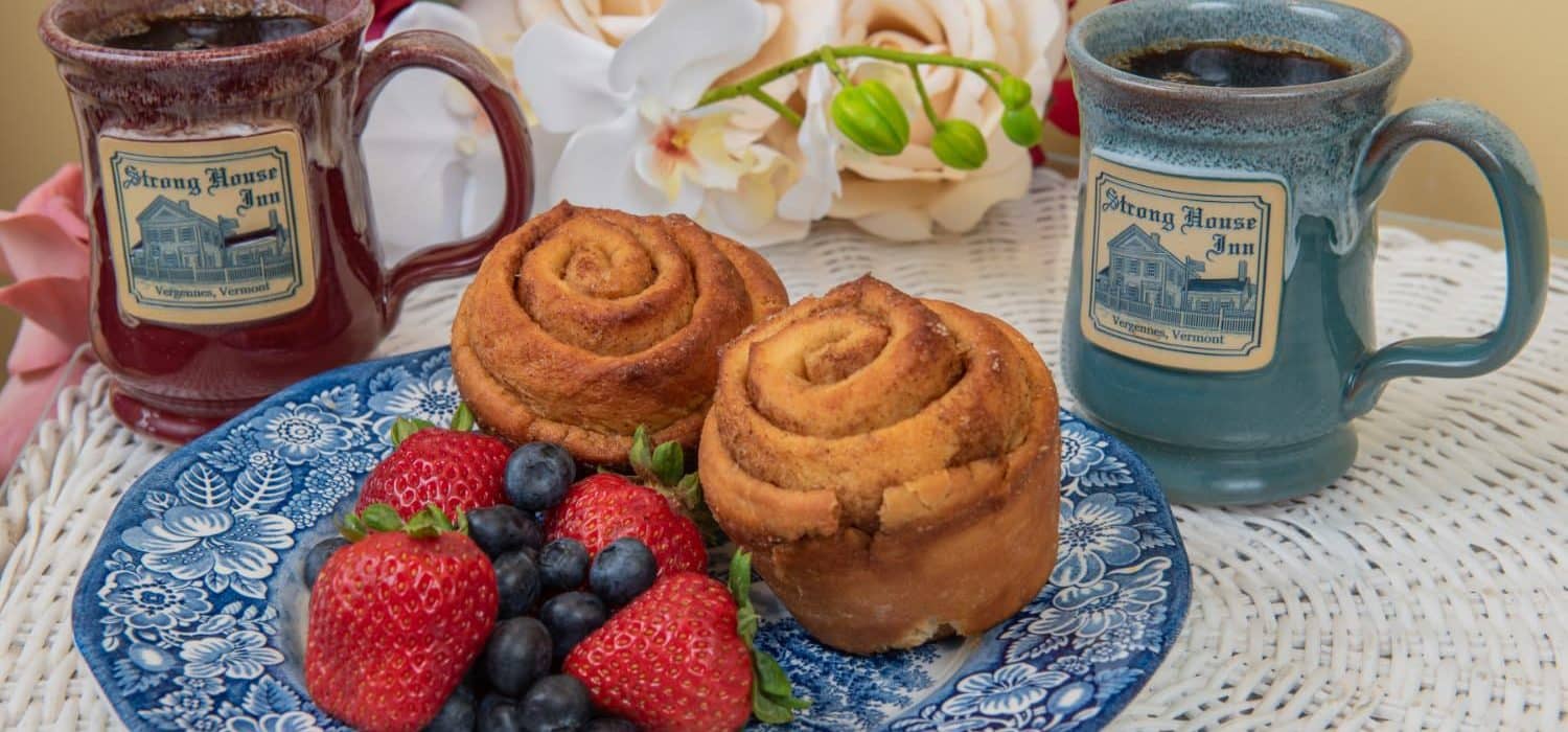 Two rolls on a blue and white plate with fruit next two two custom Deneen mugs with the Strong House Inn logo.