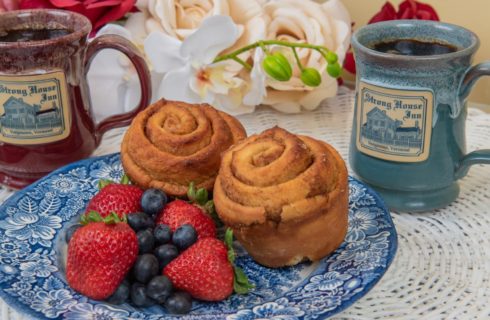 Two rolls on a blue and white plate with fruit next two two custom Deneen mugs with the Strong House Inn logo.