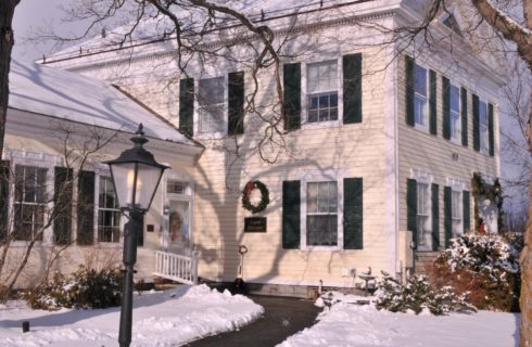 White house with wooden shutters, an iron lamp and big trees in the snow, decorated for Christmas.