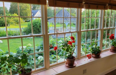 Windows with wooden blinds and red flowers in pots in sill overlook pretty garden and white gazebo.