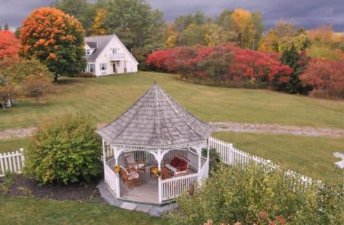 Pretty white gazebo in a green yard surrounded by glorious fall foliage backed by cloudy sly.