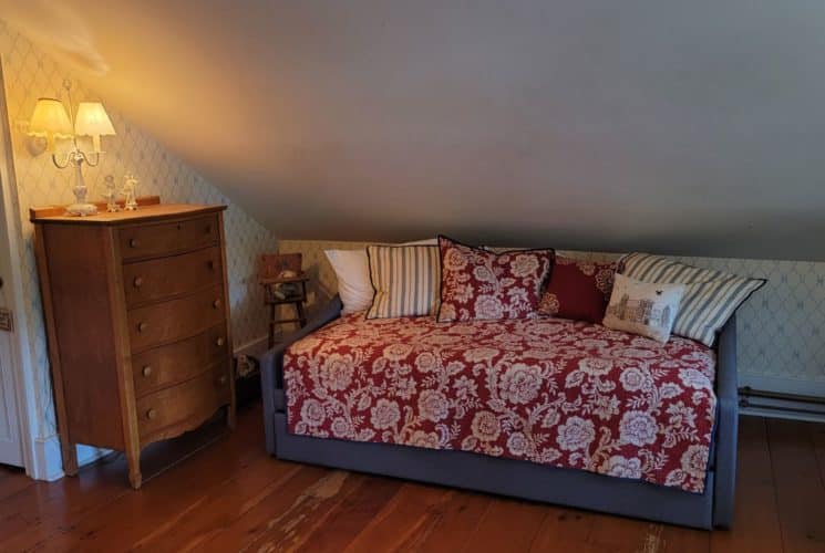 Couch-bed made up with red coverlet under dormer in bedroom with a wooden dresser.