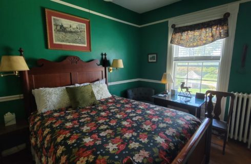 Wooden pineapple bed with flowered cover in a teal-painted bedroom with wall lights, and a table and two chairs under a window.