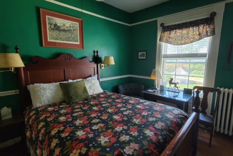 Wooden pineapple bed with flowered cover in a teal-painted bedroom with wall lights, and a table and two chairs under a window.