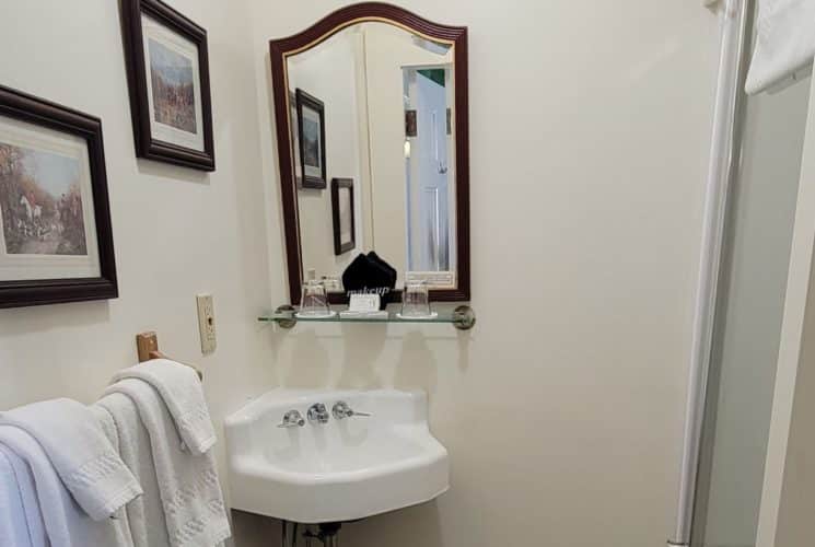 Cute bathroom with a corner sink under a mirror with white towels on a rack and two paintings.