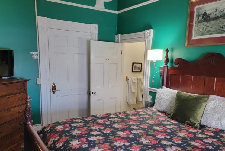 Wooden pineapple bed with flowered cover in a teal-painted bedroom with an antique door and wooden dresser with TV.