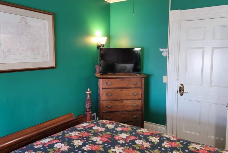 Pineapple bed with flowered cover in a teal-painted bedroom with an antique door and wooden dresser with TV.