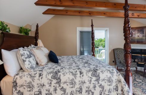 Bedroom with dormered ceiling, large cherry four-post bed, white with black bedding, Aubusson-style rug, seating area and fireplace.