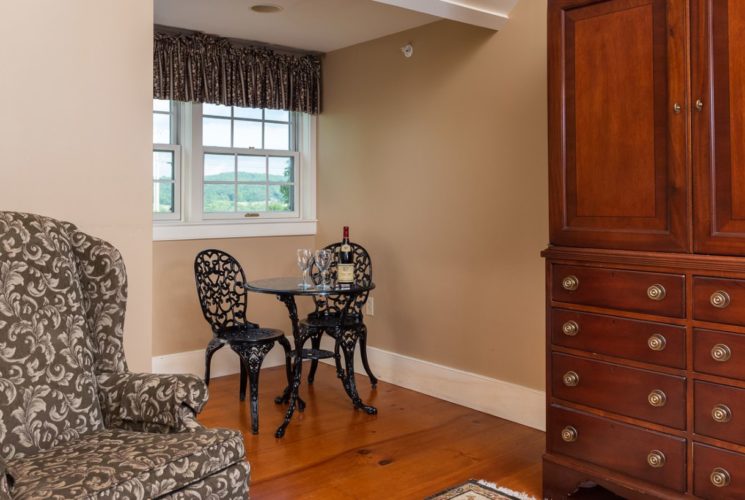 Small bistro table set with wine and glasses in corner of bedroom, overlooked by a white-painted window.