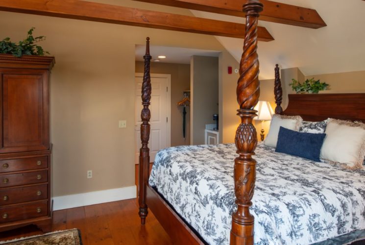Bedroom with wooden beams, large Cherrywood armoire and 4-poster bed with blue and white bedding, and wooden floor.