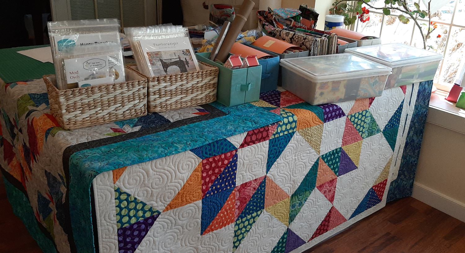 Tables covered in multicolored quilts with baskets and plastic boxes storing quilting supplies on top of the tables