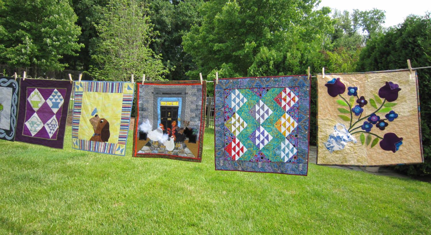 Multiple quilt squares hanging on clothes line with green grass and trees in the background