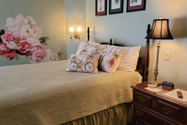 Pretty cream coverlet on wooden bed next to federal-style dresser with lamp in bedroom painted pale-green with a floral wallpaper feature.