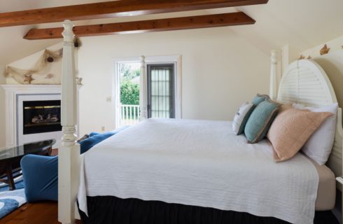 Bedroom with wooden beams, white four-poster bed with white and pastel bedding, sofa, canoe table and fireplace.