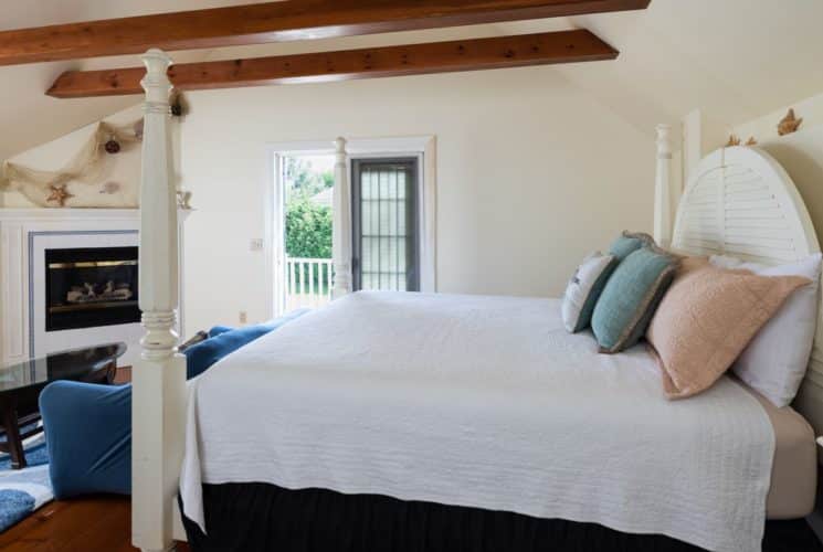 Bedroom with wooden beams, white four-poster bed with white and pastel bedding, sofa, canoe table and fireplace.