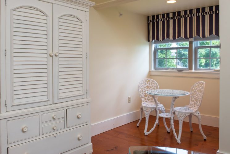 Small bistro table and chairs in corner of bedroom, overlooked by a window, next to a white wooden armoire.