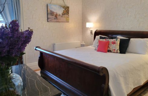 Big wooden sleigh bed with white coverlet, marble-topped table in a bedroom with beautiful wall paper and a window with blue curtains.