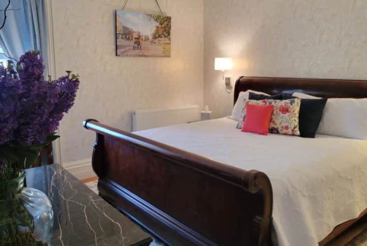 Big wooden sleigh bed with white coverlet, marble-topped table in a bedroom with beautiful wall paper and a window with blue curtains.