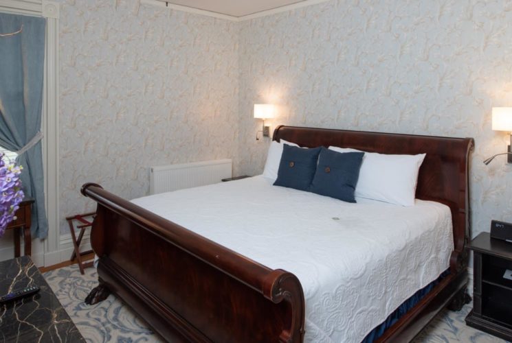 Big wooden sleigh bed with white coverlet in a bedroom with beautiful wall paper and a window with blue curtains.