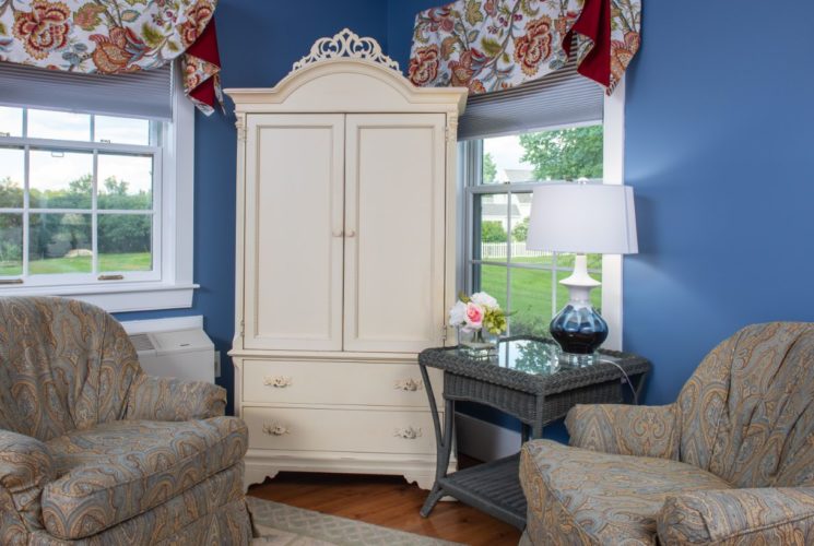 Comfy over-stuffed armchairs surround a vintage armoire and two windows in a bright bedroom painted blue.