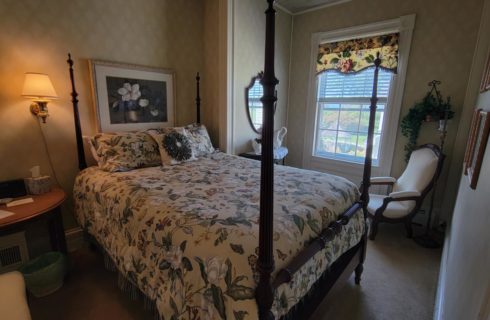 Wooden 4-post bed made up in a floral comforter in a bedroom with a cream armchair, wallpaper and a window.