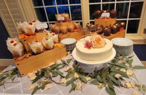 Lovely cake table at wedding reception decorated with greenery and florals holding a cake, and cupcakes on wooden stands with plates.
