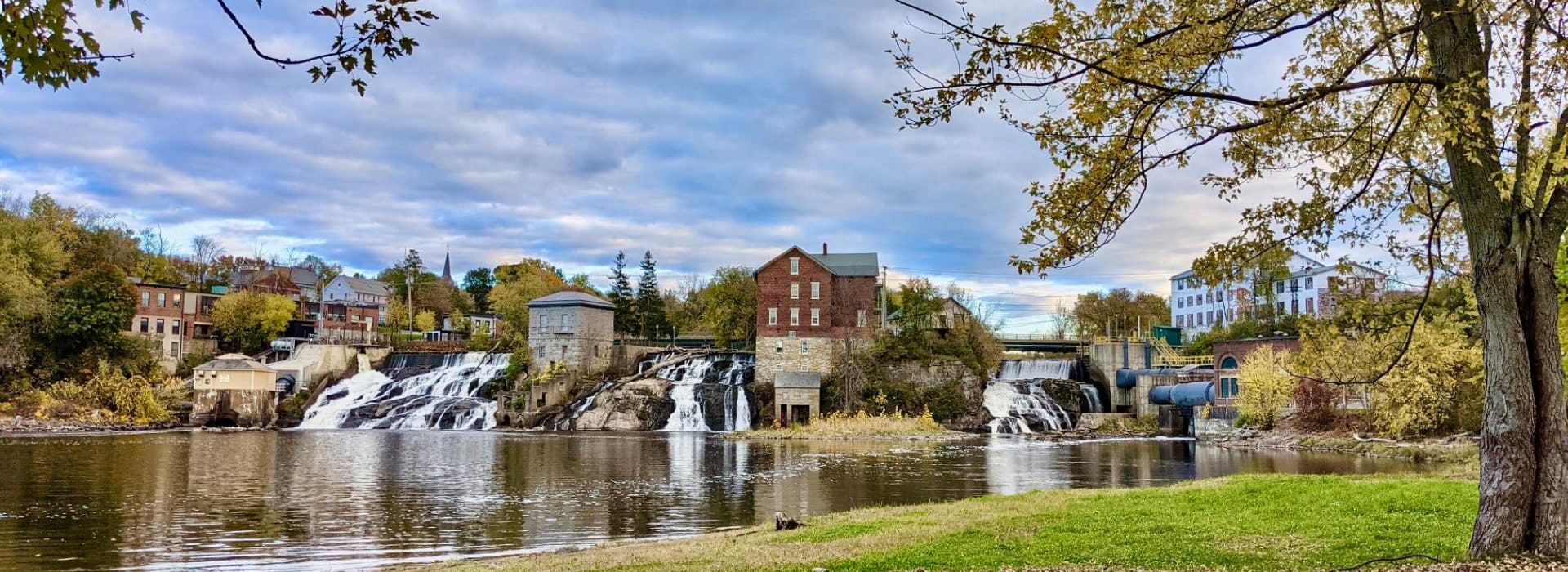 Long bridge over waterfalls separated by brick and stone buildings, surrounded by a town.