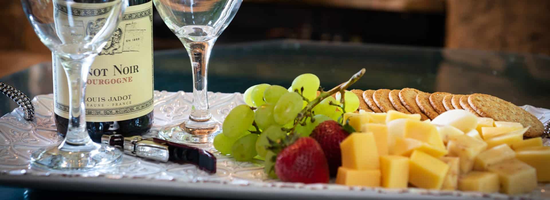 Bottle of wine and two glasses with cheese, crackers, and fruit on white ceramic tray.