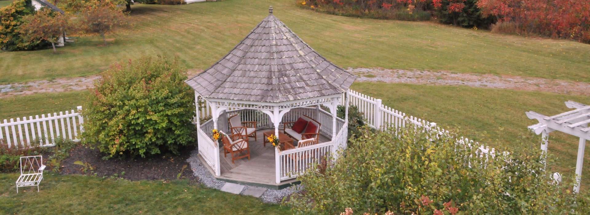 Pretty white gazebo with a grey roof covering a seating area in the corner of a yard surrounded by a white picket fence.