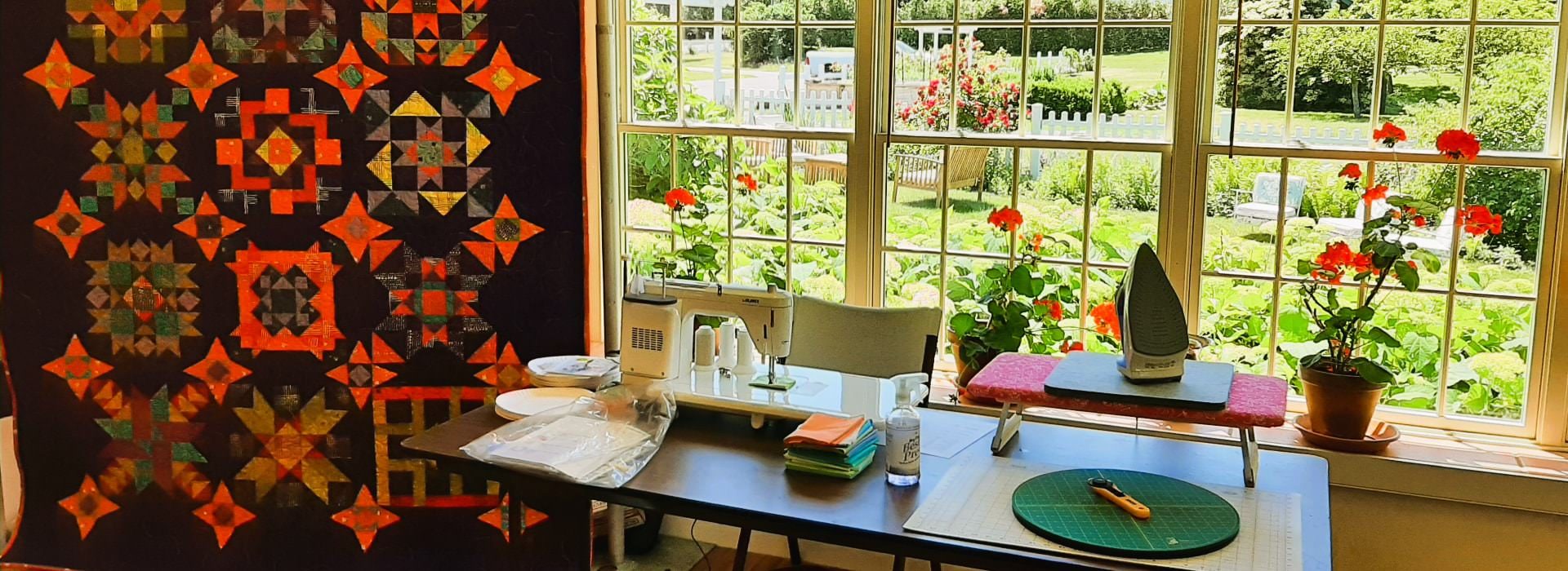 Quilt in deep colors hangs next to a table with a sewing machine and quilting supplies under a window.