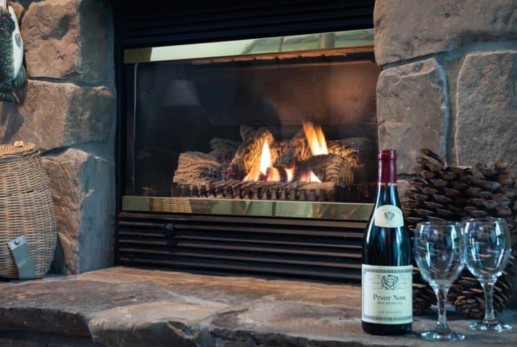 Bottle of red wine with two glasses and fishing decor on stone hearth in front of a fireplace burning merrily.