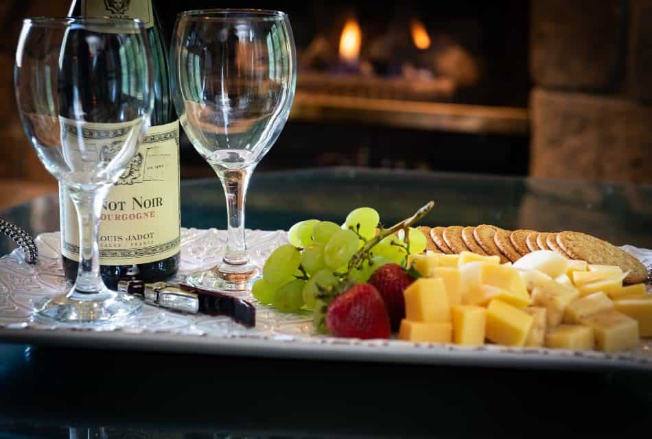 Bottle of wine and two glasses with cheese, crackers, and fruit on white tray in front of fireplace.
