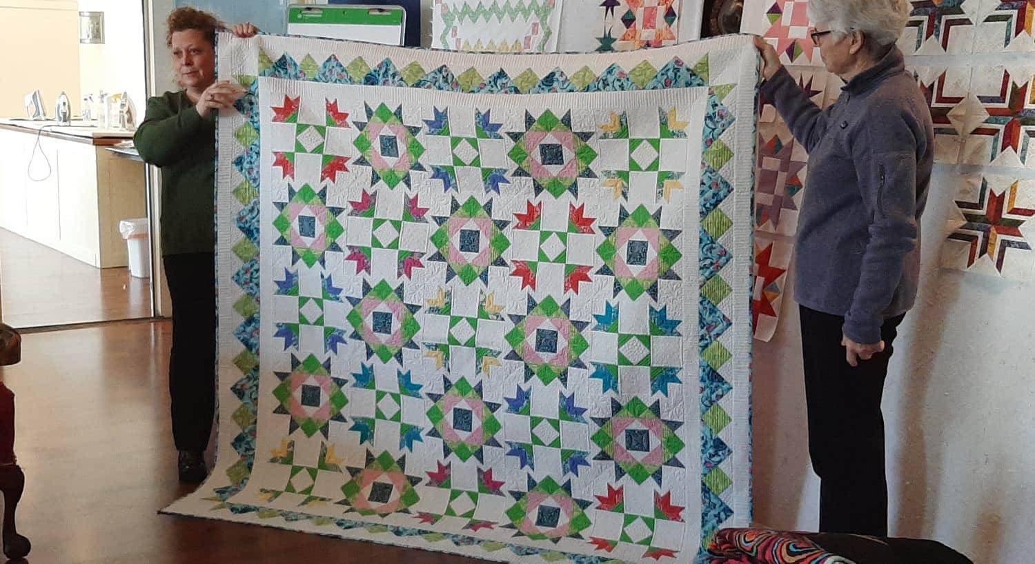 Two ladies standing and holding up a quilt with a design made up of white, green, blue, yellow, and red fabric