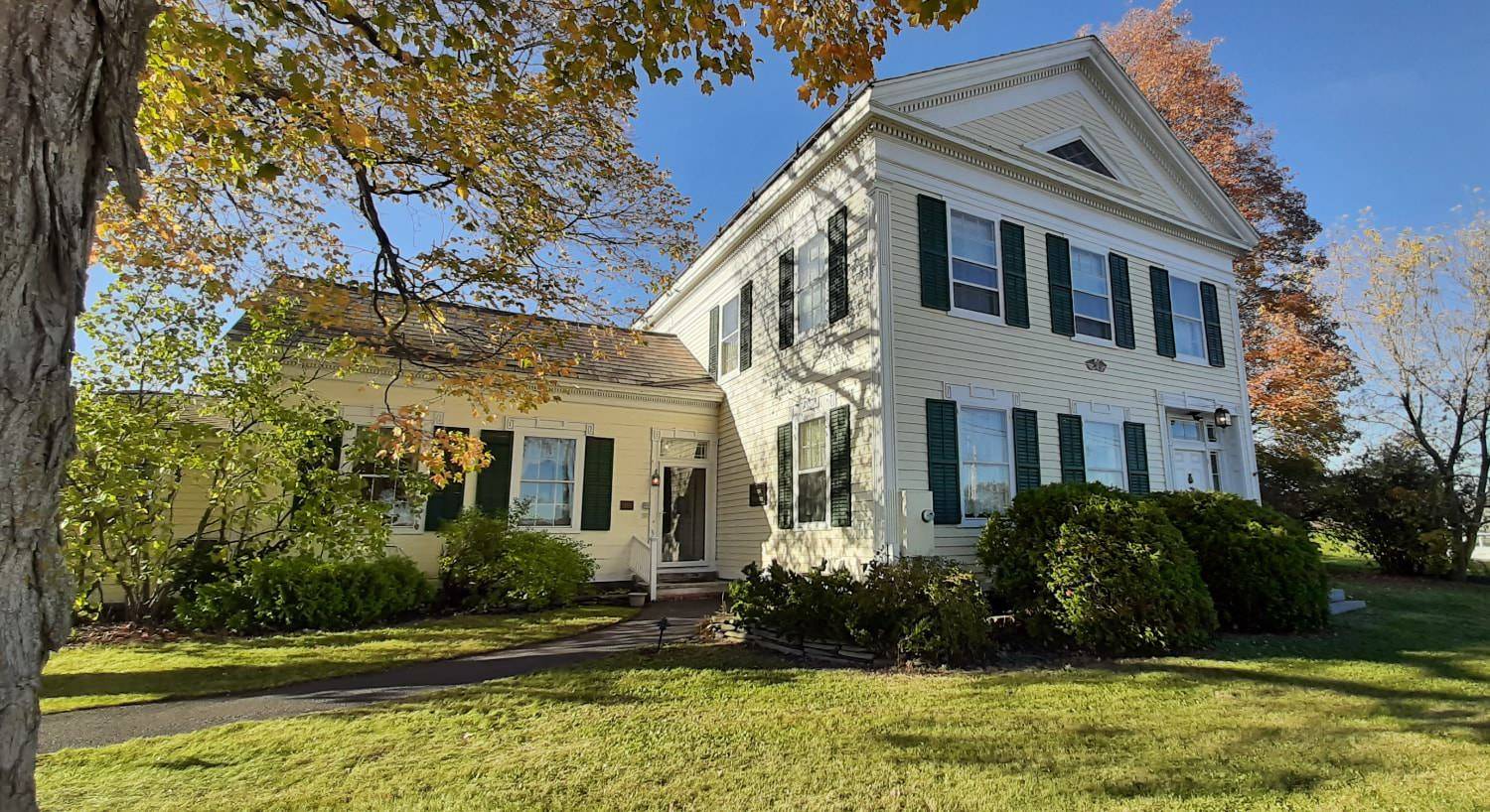 Exterior view of the property painted yellow with white trim and green shutters and surrounded by green shrubs, grass, and trees