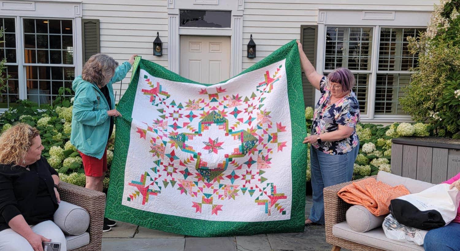 Two ladies standing and holding up a quilt with a design made up of white, green, blue, yellow, pink, and red fabric