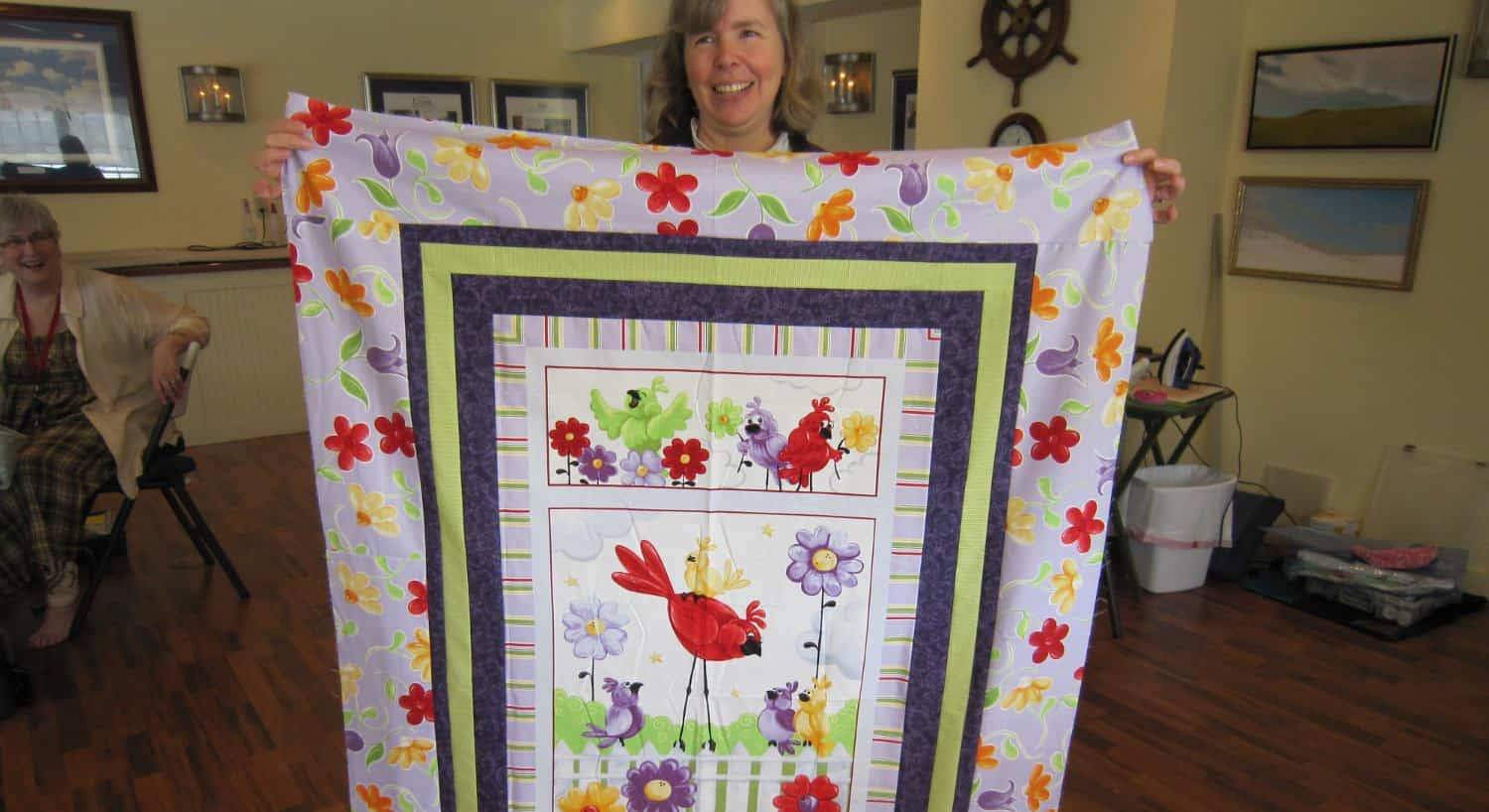 Lady holding a quilt with red, purple and yellow birds and red, purple, yellow, and orange flowers